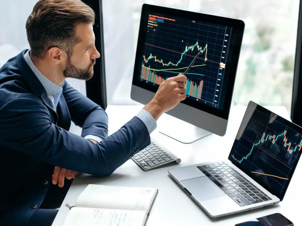 Trader señalando un gráfico de acciones en un monitor junto a una laptop